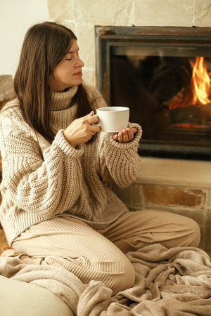 Woman in cozy sweater holding cup of warm tea at fireplace, autumn hygge. Heating house with wood burning stove. Young stylish female relaxing at fireplace in rustic farmhouseの写真素材