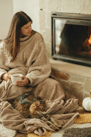 Cute cat and woman with cup of tea sitting together on cozy blanket at fireplace. Adorable tabby kitty relaxing together with owner at burning fireplace in rustic farmhouseの写真素材