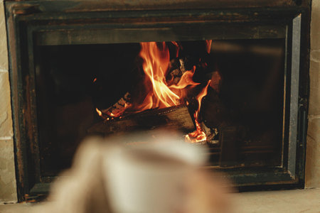Heating house with wood burning stove. Burning fireplace close up and blurred hands with cup of warm tea , autumn hygge. Relaxing and warming up at cozy rustic fireplaceの写真素材
