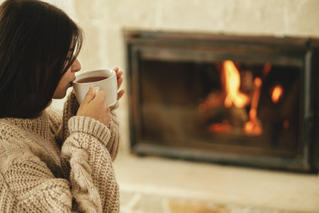 Woman in cozy sweater drinking cup of warm tea at fireplace in rustic room. Heating house in winter with wood burning stove. Young stylish female relaxing at fireplace in farmhouseの写真素材