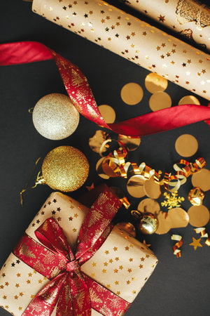 Modern christmas gift flat lay. Stylish christmas present with red ribbon, wrapping paper, golden confetti and baubles on black background. Merry Christmas and Happy Holidays!の写真素材