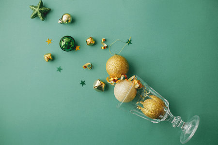 New year party concept. Stylish christmas gold baubles and confetti pouring from champagne glass on green background flat lay. Happy New year and Merry Christmas! Creative season's greeting cardの写真素材