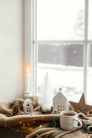 Warm cup of tea, christmas decorations, lights, little house, star on cozy blanket on windowsill. Winter hygge, Christmas still life. Cozy home on snowy day. Atmospheric scandinavian moodの写真素材