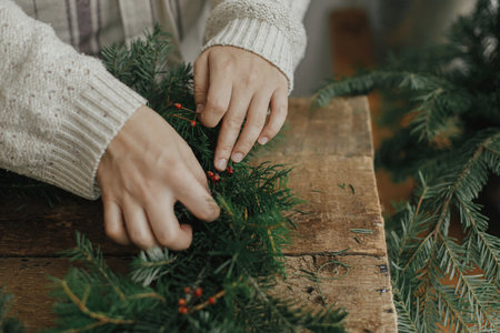 Making Christmas wreath. Woman hands holding red berries and decorating fir branches on christmas wreath on rustic wooden table. Moody holiday image. Holiday workshop, winter decorの写真素材
