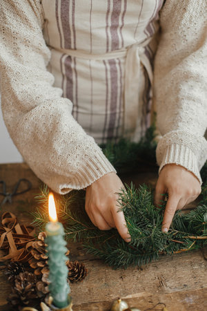 Christmas rustic wreath. Female hands holding fir branches and making wreath on rustic wooden table with ribbon, golden bells, candles. Moody Christmas image, holiday preparationsの写真素材