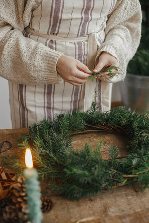 Making Christmas rustic wreath. Woman hands holding cedar branches and making wreath on rustic wooden table with ribbon, golden bells, candles. Moody holiday image. Winter holiday workshopの写真素材