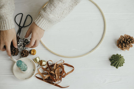 Making modern christmas wreath top view. Woman hands with round hoop, ribbon, candle, pine cones,...