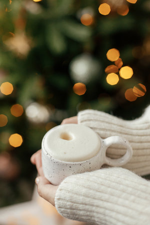 Atmospheric winter hygge. Hands holding warm cup of coffee on background of christmas tree with...