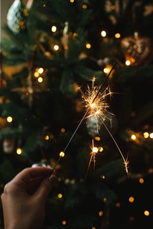 Hand holding firework against christmas tree lights in dark room. Happy New Year! Merry Christmas! Burning sparkler in female hand  on background of golden illumination bokeh. Atmospheric timeの写真素材