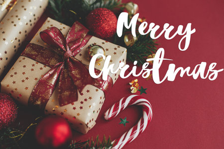 Merry Christmas text on christmas gift, wrapping paper,  fir branches, candy cane and baubles on red background. Season's greeting card. Handwritten signの写真素材