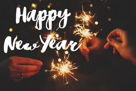 Happy New Year text sign on hands holding fireworks against christmas lights in dark room. Season's greeting card. Friends celebrating with burning sparklersの写真素材