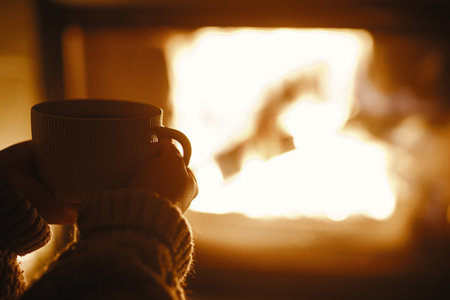 Woman holding cup of tea and warming up hands at cozy fireplace in dark evening room, close up....