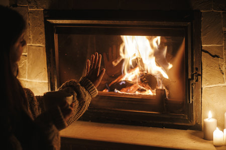 Woman hands holding cup of tea and warming up at cozy fireplace in dark evening room, close up. Fireplace heating in house, electricity blackout. Atmospheric time at fireside in homeの写真素材
