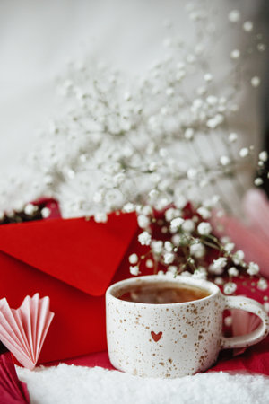 Happy valentines day! Cute stylish cup of tea with heart on cozy modern armchair with gifts, red envelope and white flowers. Valentine morning surprise for belovedの写真素材