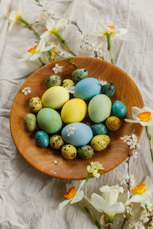 Rustic easter still life. Stylish easter eggs and blooming spring flowers in wooden bowl on linen...