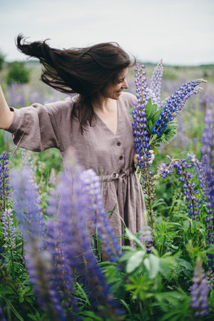 Stylish carefree woman in rustic dress holding lupine bouquet in meadow. Cottagecore aesthetics. Young female in linen dress gathering wildflowers in atmospheric summer countryside, slow lifeの写真素材