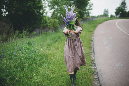 Lupine bouquet in woman hands close up in summer countryside. Cottagecore aesthetics. Young female in linen dress holding wildflowers on background of rural road and meadow, slow lifeの写真素材