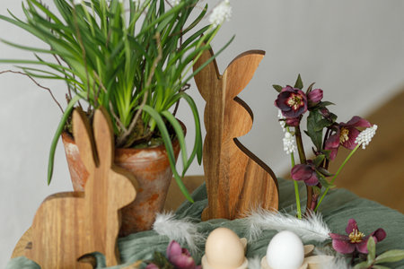 Happy Easter! Stylish wooden bunny, spring flowers, natural eggs and feathers on rustic table in room. Easter still life. Festive arrangement and decor in farmhouseの写真素材