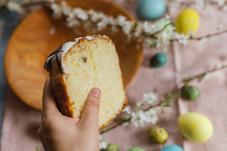 Hand holding half of baked easter cake on background of natural dyed easter eggs with spring flowers on wooden plate on rustic table.  Traditional Easter food. Happy Easter!の写真素材