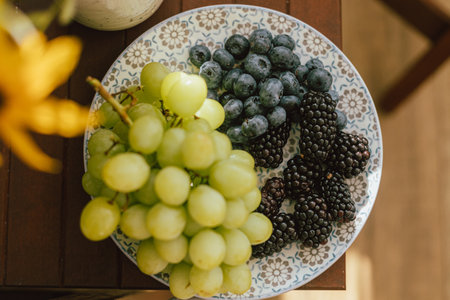 Fresh blueberries, blackberries and grapes in sunlight on ceramic plate in rustic room. healthy food aesthetics. Summer berries in light top view. Summertime in countrysideの写真素材