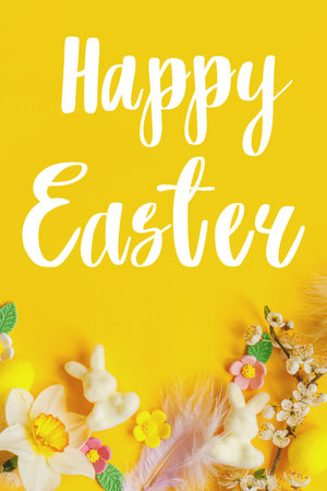 Happy Easter text sign on eggs, bunny, colorful flowers and blooming cherry petals flat lay on yellow background. Easter greeting card. Handwritten letteringの写真素材