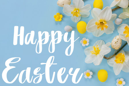 Happy Easter greeting card. Happy Easter text on stylish Easter eggs and yellow daffodils flowers flat lay on blue background. Modern Greeting card. Handwritten signの写真素材