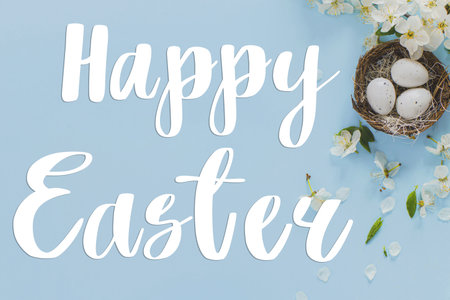 Happy Easter greeting card. Happy Easter text on Easter eggs in nest and blooming cherry petals flat lay on blue background. Modern Greeting card. Handwritten signの写真素材
