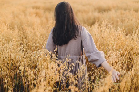 Stylish woman holding oat stems in evening light, hand close up. Rural slow life. Young female in rustic linen dress standing in harvest field in summer countryside. Atmospheric tranquil momentの写真素材