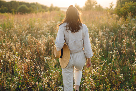 Stylish boho woman with straw hat walking among wildflowers in warm sunset light. Atmospheric moment. Summer delight and travel. Young female in rustic linen cloth relaxing in summer meadow.の写真素材