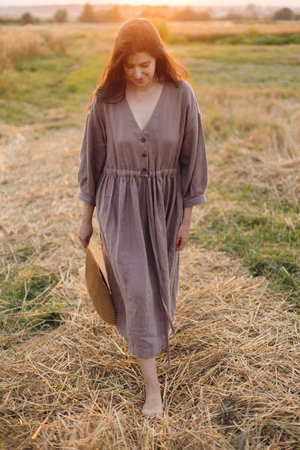 Stylish woman with straw hat walking at oat field in sunset light. Atmospheric tranquil moment. Young female in rustic linen dress relaxing in evening summer countryside, rural slow lifeの写真素材