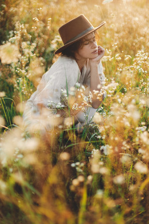 Stylish boho woman in hat sitting among wildflowers in warm sunset light. Summer delight and travel. Young female in rustic linen cloth relaxing in summer meadow. Atmospheric momentの写真素材