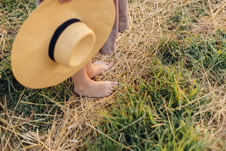 Woman with hat in hand walking barefoot on straw field in sunset light, close up. Rural slow life. Atmospheric tranquil moment. Young female in rustic linen dress relaxing in summer countrysideの写真素材