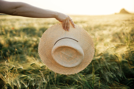 Straw hat in woman hand in sunset light on background of barley field. Stylish female enjoying evening summer countryside. Atmospheric tranquil moment, rustic slow lifeの写真素材