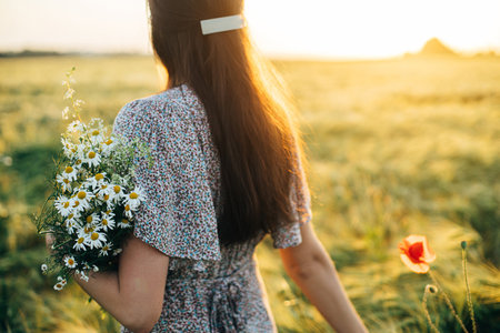 Beautiful woman with wilflowers bouquet standing in barley field in sunset light. Stylish female relaxing in evening summer countryside and gathering flowers. Atmospheric tranquil momentの写真素材