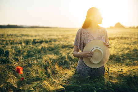 Beautiful woman in floral dress standing in barley field in sunset light. Atmospheric tranquil moment, rustic slow life. Stylish female holding straw hat and enjoying evening summer countrysideの写真素材