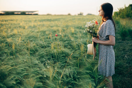 Beautiful woman with straw hat and wildflowers enjoying sunset in barley field. Atmospheric tranquil moment, slow life. Stylish female gathering flowers and walking in evening summer countrysideの写真素材