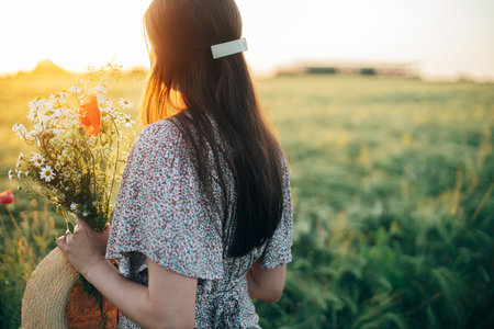 Beautiful woman with wildflowers enjoying sunset in barley field. Atmospheric tranquil moment, rustic slow life. Stylish female gathering flowers and relaxing in evening summer countrysideの写真素材