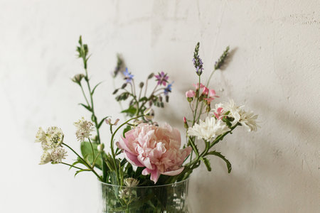 Stylish bouquet with peony and wildflowers on tile shelf on rustic wall background in modern room. Beautiful flowers in glass vase gathered from garden, summer floral arrangement in home.の写真素材