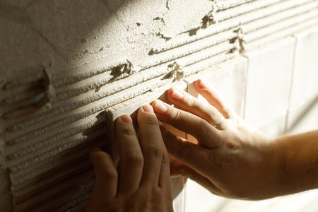 Hands laying modern square tile on adhesive close up. Worker installing stylish white tiles on plaster wall in sunlight. Renovationの写真素材