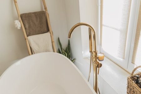 Stylish bathroom interior. Modern bathtub with golden faucet from floor, wooden ladder with towels...
