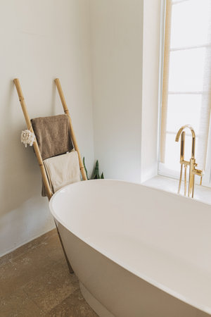 Stylish boho bathroom design. Modern bathtub with golden faucet from floor, wooden ladder with towels and big window, modern eco bathroom interior.の写真素材