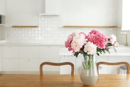 Beautiful peonies in vase on wooden table on background of stylish white kitchen with island, wooden shelves and appliances in new house. Modern kitchen interior in farmhouseの写真素材