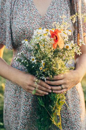 Beautiful woman holding wildflowers bouquet close up in sunset light in barley field. Stylish female relaxing in evening summer countryside and gathering flowers. Atmospheric tranquil momentの写真素材
