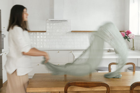 Blurred woman arranging green textile table runner on wooden table in new modern home. Motion image of young female decorating dining room on background of minimal kitchen, housekeepingの写真素材