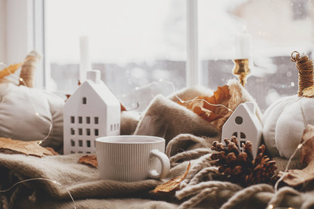 Autumn still life. Warm cup of tea, cozy pumpkin pillows, fall leaves, candle, lights and cute buildings decoration on brown scarf on windowsill. Hygge fall home decor. Happy Thanksgivingの写真素材