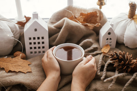 Cozy Autumn. Hands holding warm cup of tea with stylish pumpkin pillows, fall leaves, lights and cute buildings decor on brown scarf on windowsill. Autumn hygge, fall home decorの写真素材