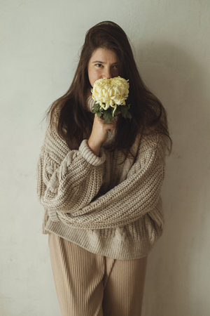Stylish woman in cozy knitted sweater posing with chrysanthemum flower on background of rural authentic wall. Moody fall. Young fashionable female in hygge autumn homeの写真素材