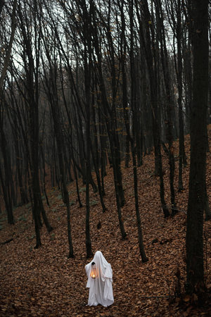 Spooky ghost holding glowing lantern in moody dark autumn forest. Person dressed in white sheet as ghost with light in evening fall woods. Happy Halloween! Trick or treatの写真素材
