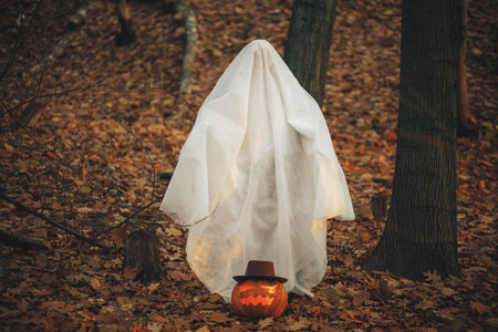 Stylish ghost and jack o lantern in moody autumn forest. Person dressed in white sheet as ghost with pumpkin standing in evening fall woods. Happy Halloween! Trick or treatの写真素材