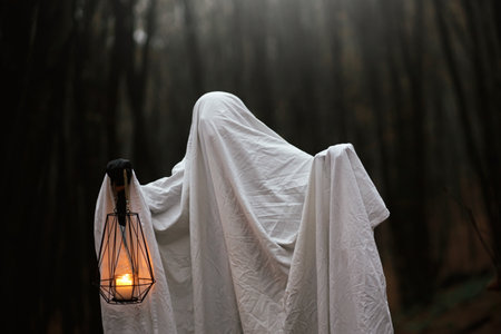 Happy Halloween! Spooky ghost holding glowing lantern in moody dark autumn forest. Person dressed in white sheet as ghost with light in evening fall woods. Boo! Horror timeの写真素材